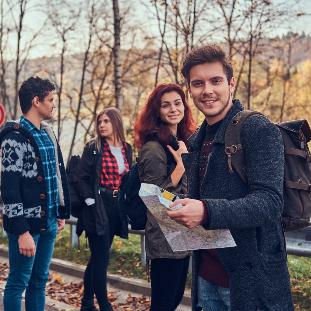 Travel, hitchhiking, adventure concept. Group of young hikers standing on the road sidelines at beautiful autumn forest, guy with map planning hike.